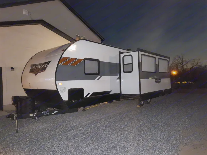Nighttime rv rental: A Wildwood travel trailer with an open slide-out is parked on gravel next to a building.. Forest River Wildwood 2021