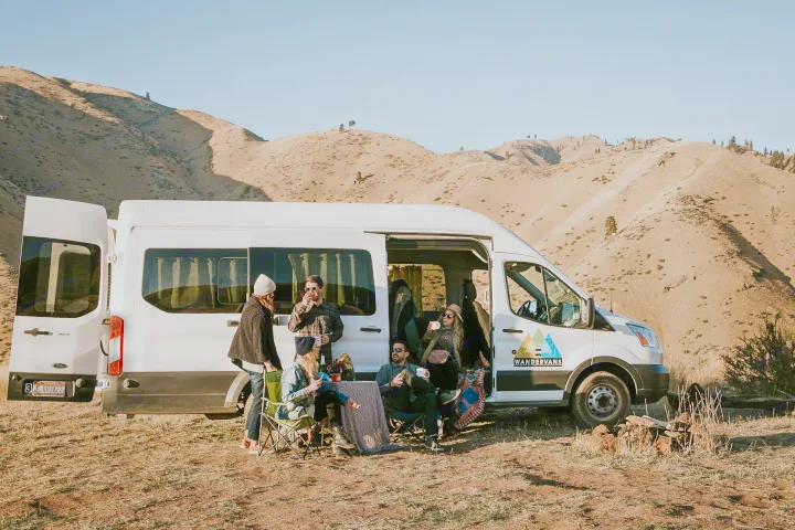 A group socializes by an rv rental camper van amidst dry mountains, enjoying an outdoor adventure.. Ford Transit 350 2021