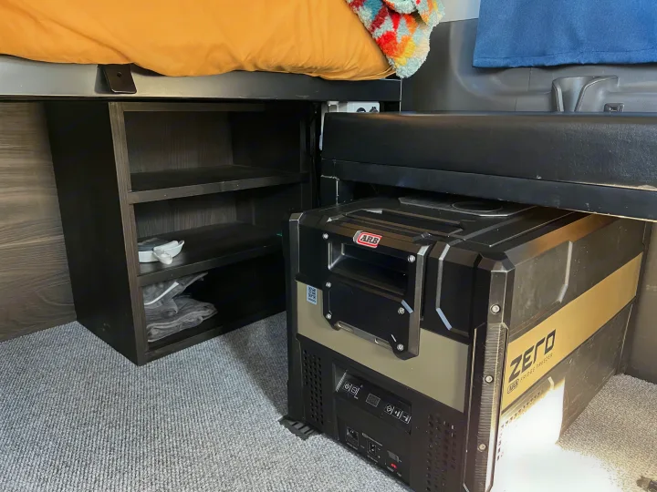 RV rental interior: smart setup includes a raised bed, dark storage shelves, and an ARB ZERO portable fridge/freezer.. Ford transit 2019