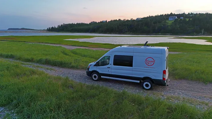 Explore scenic routes with an RV rental. White Casual camper van on a gravel path by water and hills at dusk.. Ford Transit 2020