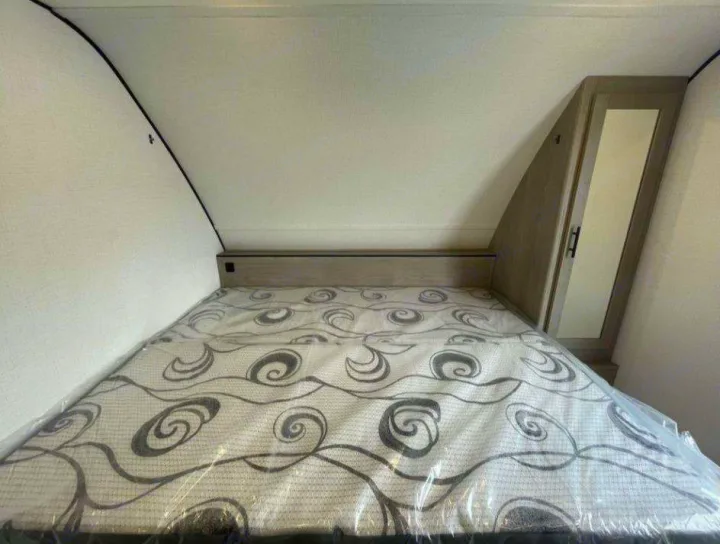 Compact RV rental bedroom featuring a new, plastic-covered mattress and angled built-in storage cabinet.. Dutchmen Coleman 1905BH 2024
