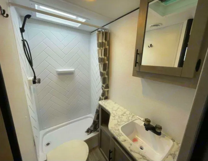 Inside a clean RV rental: compact bathroom with herringbone shower, toilet, sink, and mirrored medicine cabinet.. Dutchmen Coleman 1905BH 2024