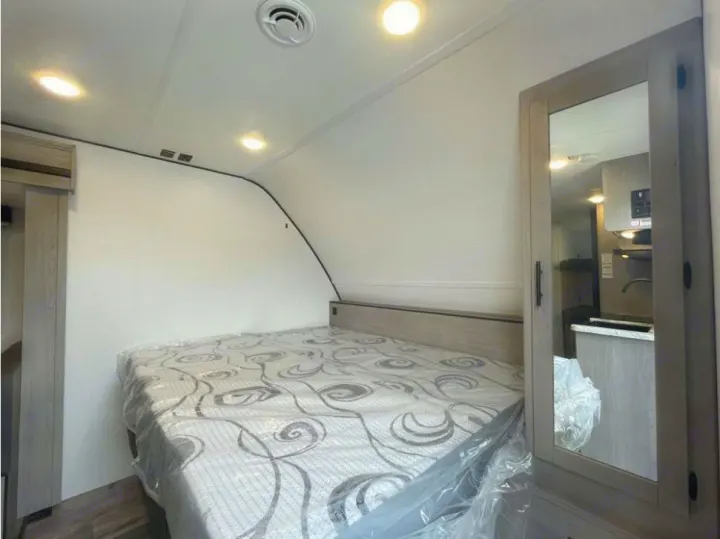 Clean RV rental bedroom featuring a bed with a fresh mattress. Mirrored closet reflects the modern kitchen space.. Dutchmen Coleman 1905BH 2024
