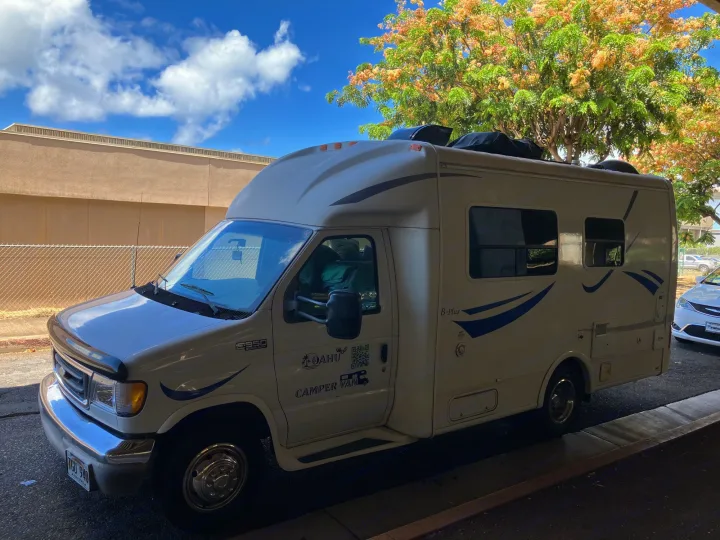 White Ford E350 Super Duty RV rental, an Oahu camper van, parked under a sunny sky with a flowering tree.. Ford Econoline 2004