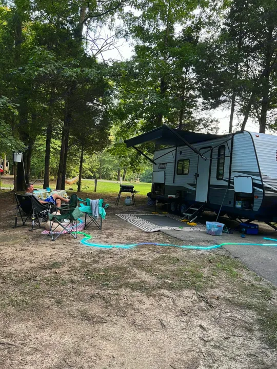 An RV rental campsite setup, featuring a man relaxing by his Coleman travel trailer with awning, dog, and grill. Shady trees overhead.. Dutchmen Coleman Lantern LT 2021