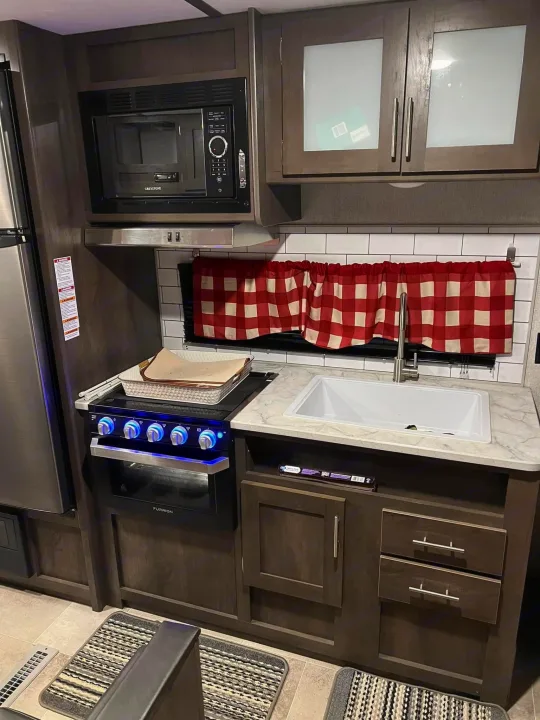 This RV rental features a compact kitchen with a microwave, stove with blue lights, sink, and checkered curtains.. Dutchmen Coleman Lantern LT 2021