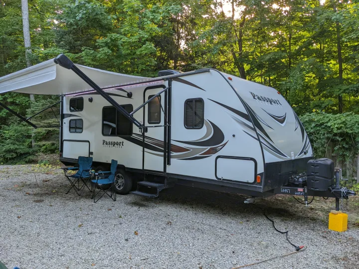 An RV rental: a Passport Ultra-Lite travel trailer with an extended awning and chairs on a gravel site, nestled among trees.. Keystone RV Passport Ultra Lite 2018