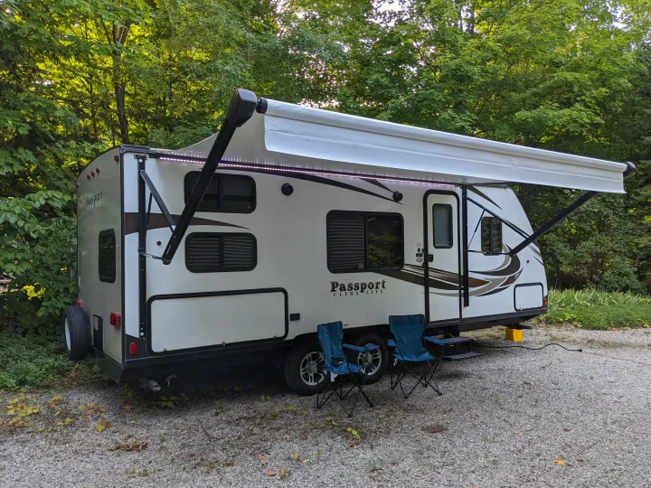 Cozy RV rental parked on a gravel campsite with an extended awning and chairs, surrounded by lush green trees.. Keystone RV Passport Ultra Lite 2018