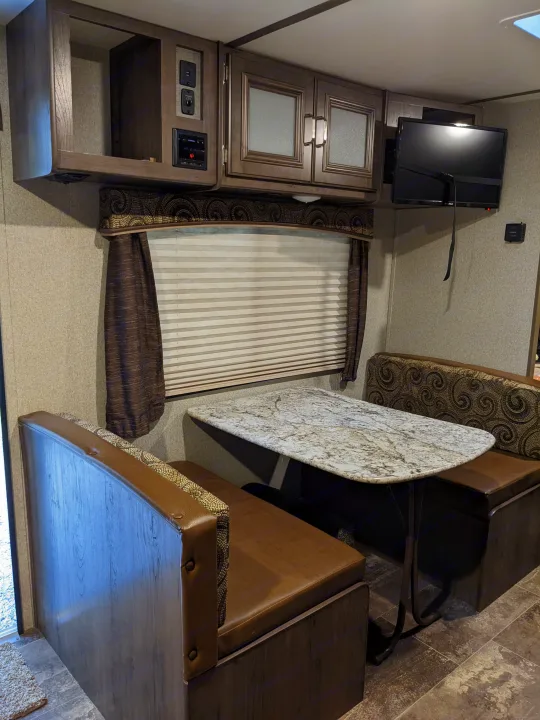 Inside this RV rental: a cozy dinette with a faux marble table, brown bench seats, overhead cabinets, and a wall-mounted TV.. Keystone RV Passport Ultra Lite 2018