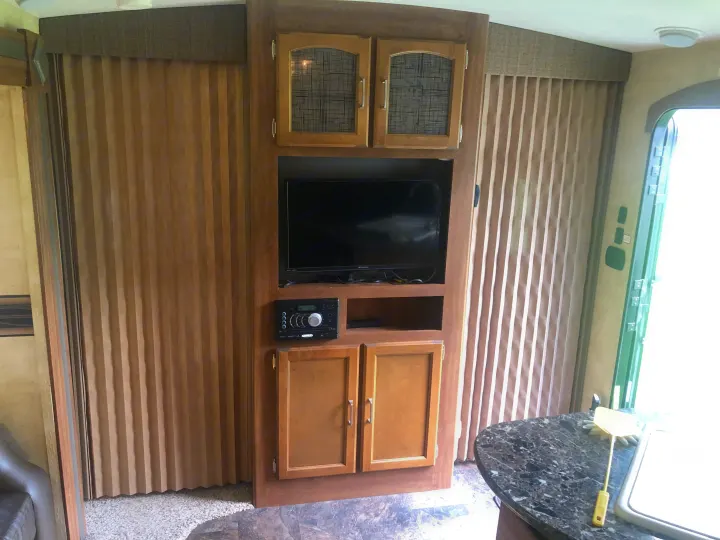 RV rental interior with an entertainment center featuring a TV, stereo, and wooden cabinets, alongside accordion blinds.. Keystone RV Springdale 2017