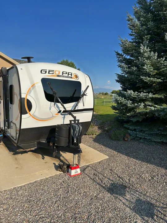 A white GEO PRO RV with a front bike rack, parked on gravel next to a tree and field. A potential rv rental for outdoor adventures.. Forest River GeoPro 2022