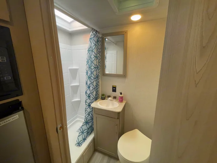 Step inside this RV rental bathroom with a white shower stall, blue curtain, compact sink, mirror, and toilet for comfortable camping.. Keystone Springdale 1800BH 2024