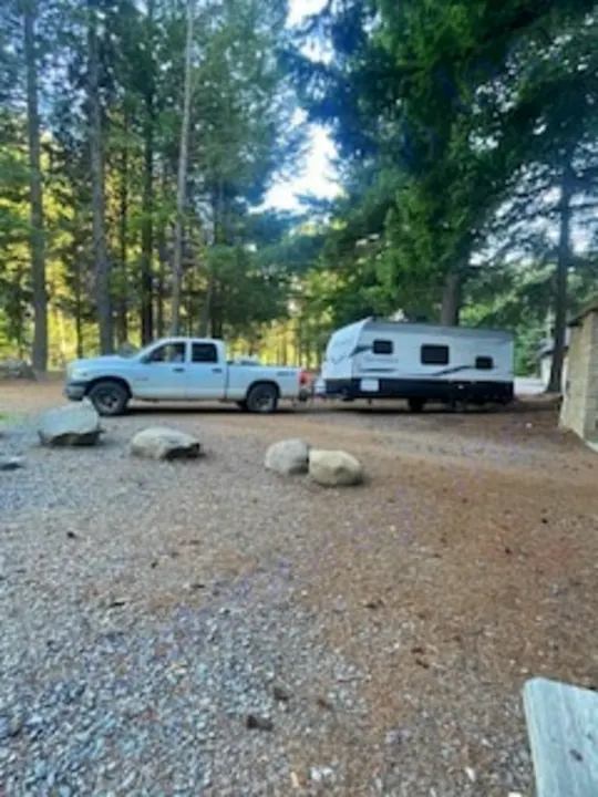 A white pickup truck tows an RV rental travel trailer, parked on gravel amidst tall trees, ready for a camping adventure.. Keystone RV Springdale 2022