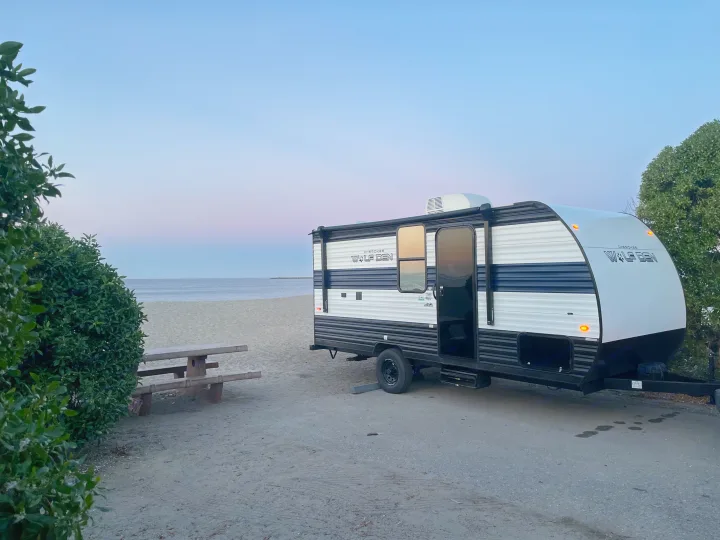 RV rental by the beach: a white and blue travel trailer parked on sand near a picnic table with ocean and sunset sky.. Forest River Wolf Den 16EV 2024