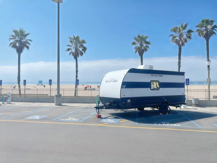 This rv rental, a Wolf Den travel trailer, is parked in an accessible lot by a sunny beach with palm trees.. Forest River Wolf Den 16EV 2024
