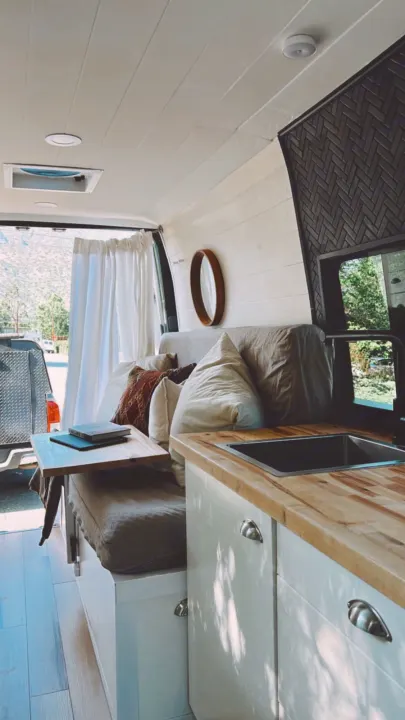 Bright RV rental interior: soft cushions, small kitchen, wooden table, and scenic mountain view from open back.. Mercedes-Benz Sprinter 2016