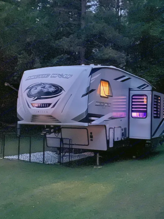 An illuminated white Arctic Wolf fifth-wheel RV rental is parked on grass at dusk, with warm and purple interior lights on.. Forest River Cherokee Arctic Wolf 2021