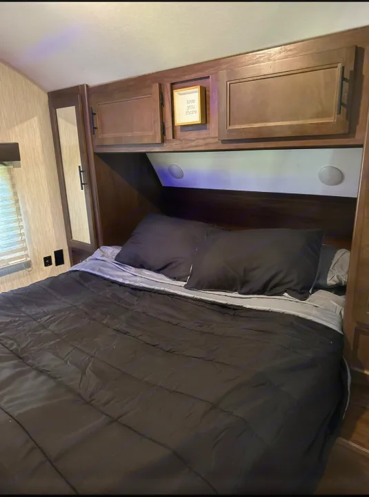 Cozy RV rental bedroom featuring a dark-themed bed with pillows, overhead wooden cabinets, and a side window.. Forest River Cherokee Grey Wolf 2021