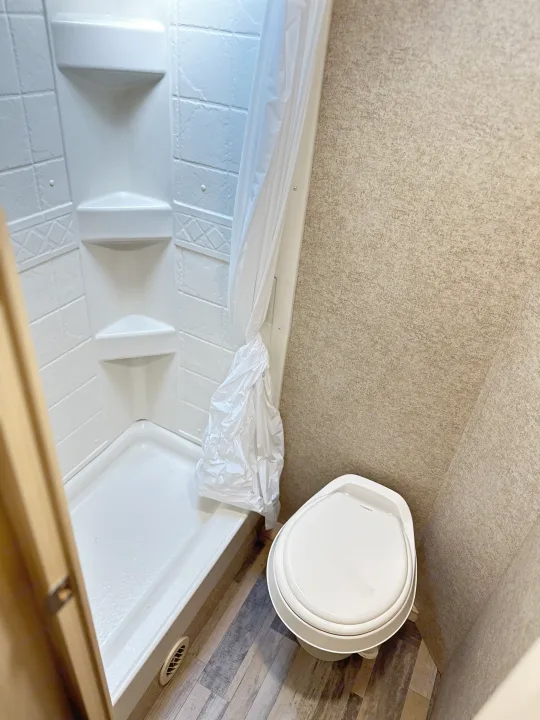 An RV rental bathroom interior, showing a white corner shower with curtain and a Dometic toilet on wood-look floor.. Minnie Micro Minnie 2018
