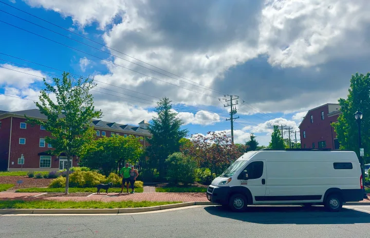 A white RV rental van sits on a street with a couple and their dog on a sidewalk. Buildings and trees frame the background under a cloudy sky.. Custom Ram Promaster 2021