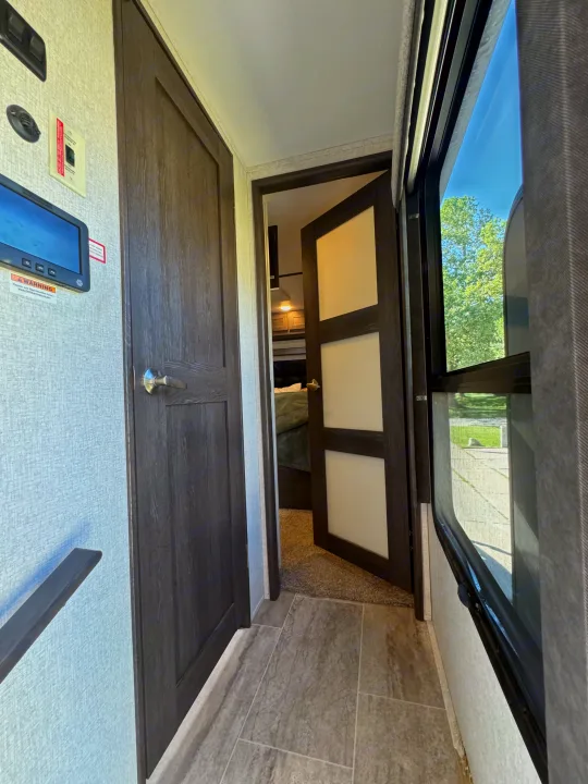 RV rental interior: compact passage with a control panel, dark wood doors, and window, leading to a private bedroom.. Keystone Raptor 2020
