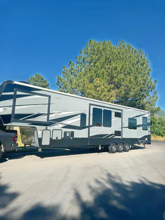 Large gray Raptor Fifth Wheel RV rental parked with a truck under a blue sky, ideal for trips and camping.. Keystone Raptor 2020