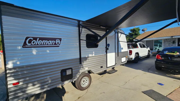 Coleman travel trailer with awning extended, parked on a paved driveway next to a pickup truck. Consider this for RV rental!. Coleman 17b 2024