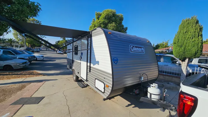 Bright blue sky over a parked Coleman travel trailer with its awning out, ideal for your next RV rental trip.. Coleman 17b 2024