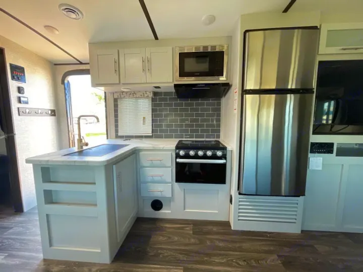 Modern RV rental kitchen featuring a stainless fridge, oven, microwave, sink, and light gray cabinetry. Bright and compact for travel.. Keystone RV Bullet Ultra Lite 2021