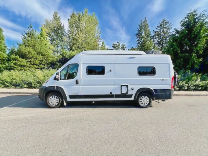 A white Winnebago Solis RV rental camper van parked on asphalt with trees and blue sky. Ready for adventure.. Winnebago Winnebago 2023