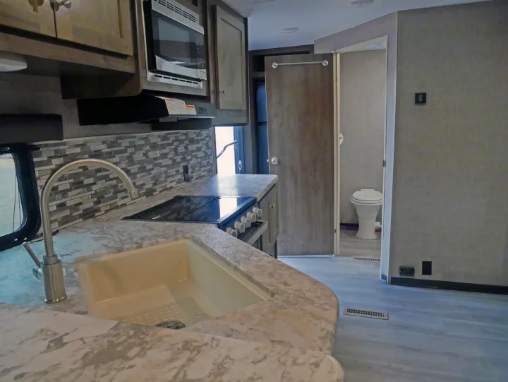Modern RV rental kitchen featuring a sink, stove, microwave, and a glimpse into the attached bathroom.. Dutchmen Aspen Trail 2020