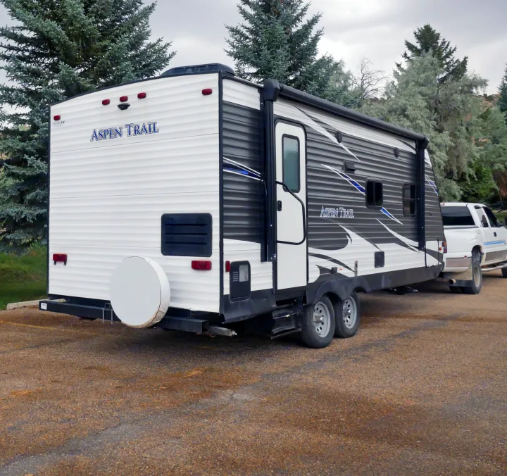 An Aspen Trail RV rental travel trailer is hitched to a white dually pickup truck, ready for camping. Trees and a cloudy sky surround them.. Dutchmen Aspen Trail 2020