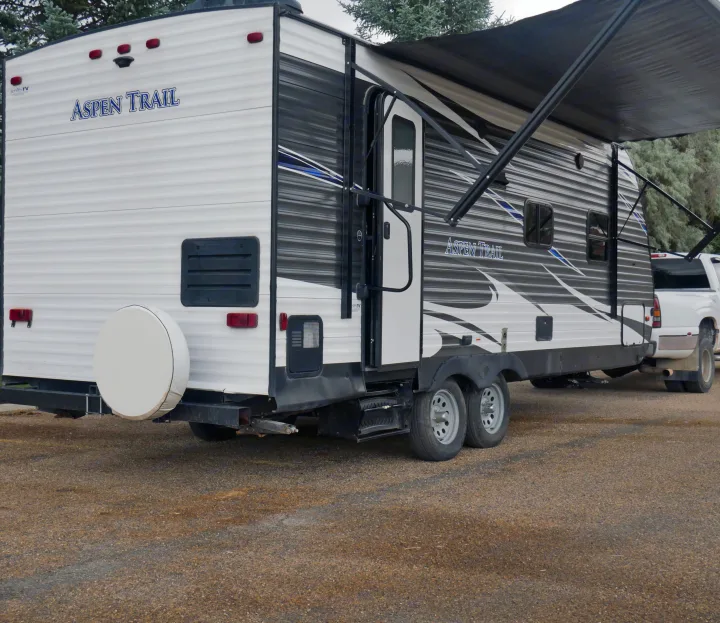 White and gray Aspen Trail travel trailer (an rv rental) parked on gravel, hooked to a pickup, with its awning out.. Dutchmen Aspen Trail 2020