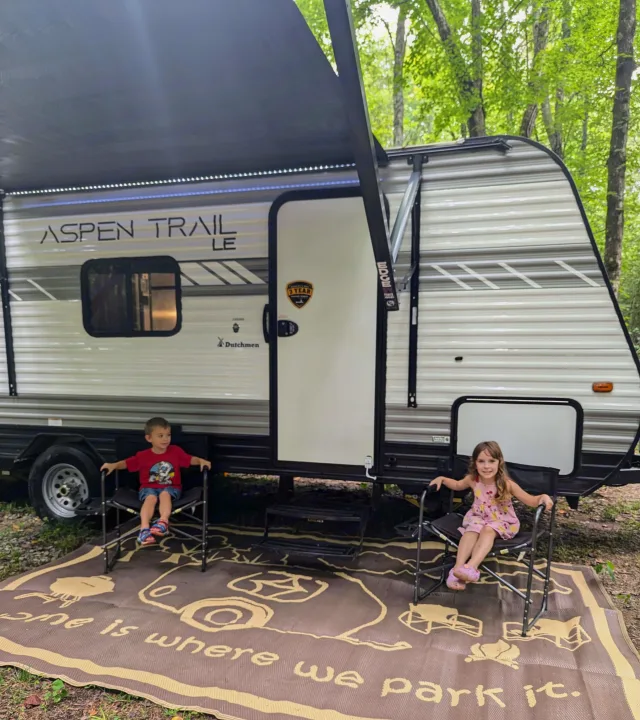 An RV rental offers cozy camping! Two kids relax on a "Home is where we park it" mat by their travel trailer.. Aspen Trail LE 2022
