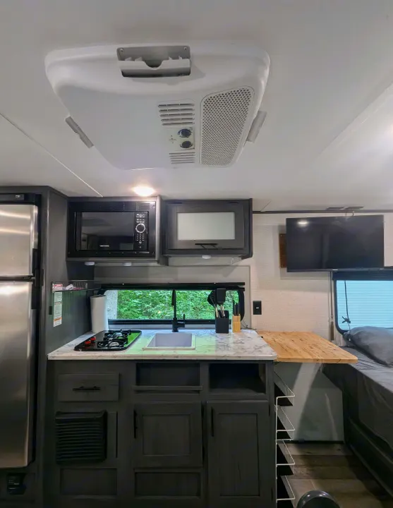 Modern rv rental interior showing a compact kitchen area with appliances, sink, and adjacent TV and bed.. Aspen Trail LE 2022