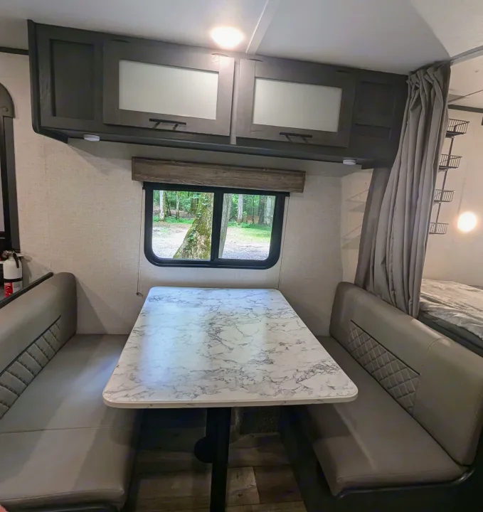 Inside this RV rental: a comfy dinette with gray benches, marble-look table, and a window revealing a forest view.. Aspen Trail LE 2022