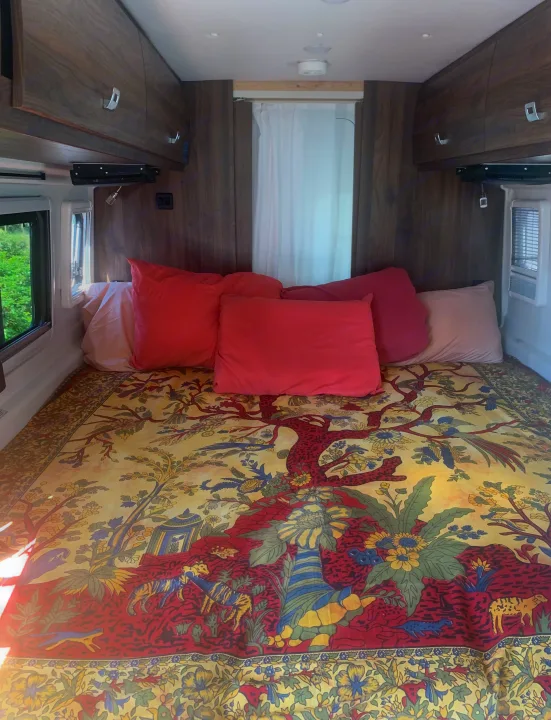 Inside an RV rental: a vibrant bed with an ornate tapestry, plush pillows, and built-in wood cabinets.. Winnebago Travato 2022