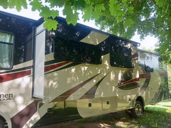 An RV rental in cream, brown, and maroon, showcasing its extended slide-out and side storage compartments, parked under green trees.. Forest River Coachmen Mirada 2018