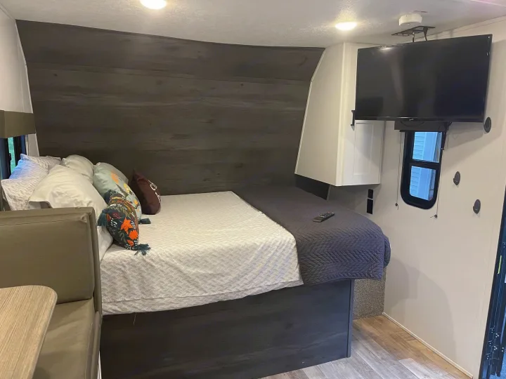 RV rental: A compact bedroom interior featuring a large bed with pillows, dark wood paneling, a wall-mounted TV, and a dinette.. Keystone RV Hideout 2021