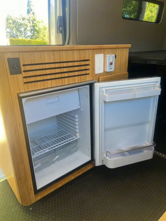 An RV rental interior with a built-in, open refrigerator/freezer in bamboo cabinetry, plus power outlets & a monitor.. Mercedes Sprinter Sprinter 2500 2021