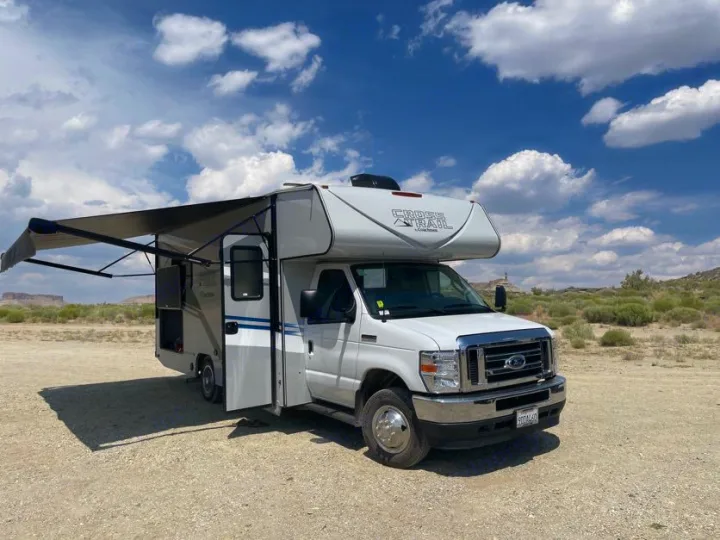 A white Class C RV rental with its awning extended is parked on a dirt lot, ready for adventure in a scenic desert landscape.. Forest River Coachmen Crosstrail 2023