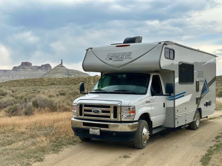 Explore the desert! This RV rental is parked on a dirt road, facing unique rock formations and mountains under a cloudy sky.. Forest River Coachmen Crosstrail 2023