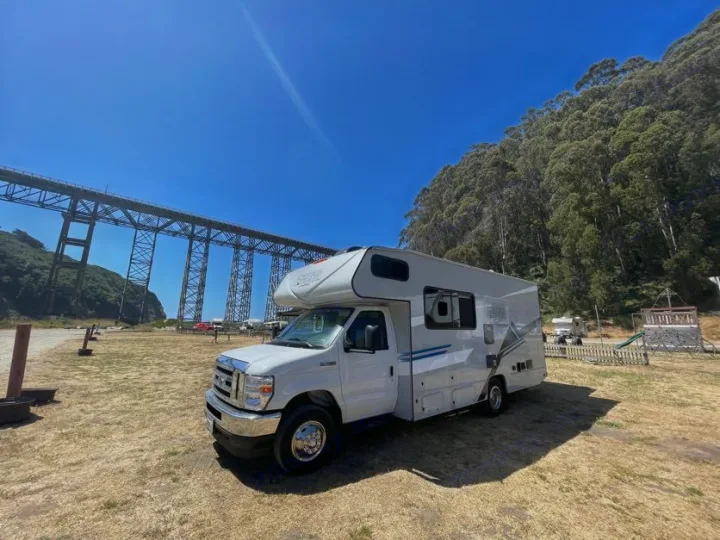 RV rental at a scenic spot. A massive steel bridge spans the left, dense forest covers the right hill.. Forest River Coachmen Crosstrail 2023