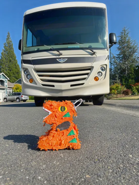 An orange dinosaur piñata faces a large, beige RV rental on asphalt. Ready for adventure and a fun road trip!. Flair-Fleetwood Flair-Fleetwood Motorhome 2017