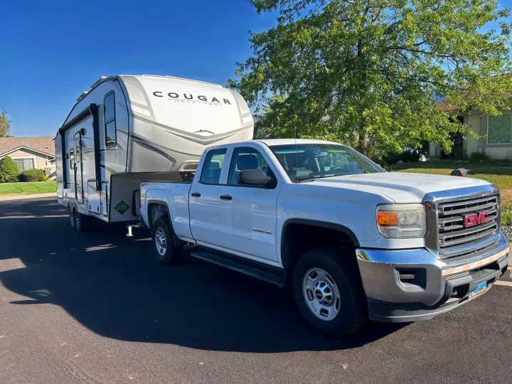 White GMC truck pulling a Cougar Half-Ton fifth-wheel RV on a sunny day. Great for an RV rental or camping trip.. Keystone RV Cougar 2023