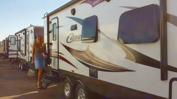 A woman stands on steps of a Cougar travel trailer. Many RVs are lined up at an outdoor RV rental lot.. Keystone RV Cougar 2015