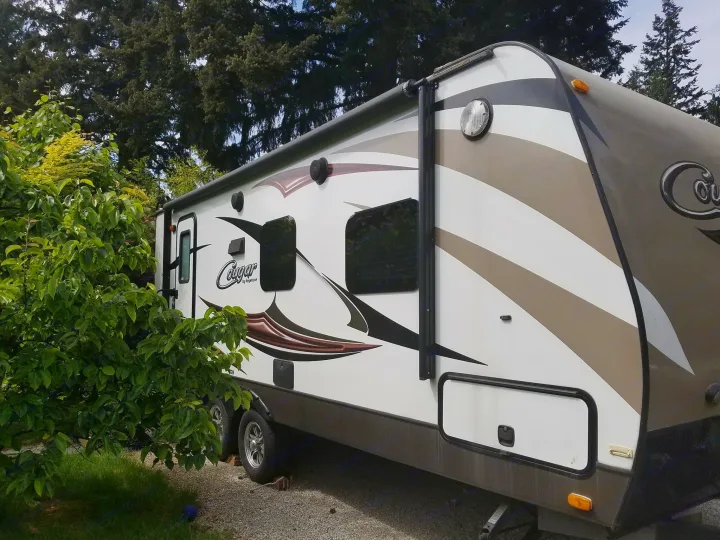 Side view of a white and brown Cougar by Keystone RV rental, parked on gravel near green trees, ready for a trip.. Keystone RV Cougar 2015