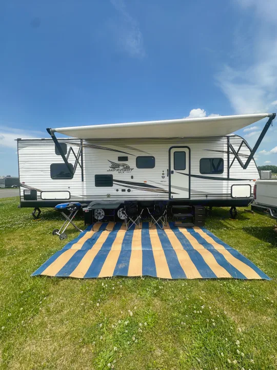 RV rental set up at a sunny campsite with an awning, mat, and grill on green grass under a blue sky.. Jayco Jay Flight SLX 2019