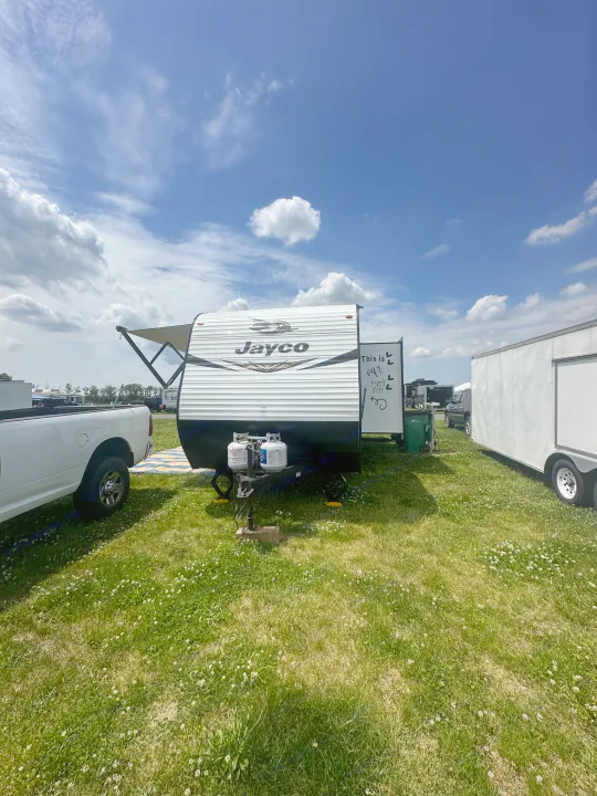 Jayco RV rental parked on a sunny, green field with a white truck & trailer nearby. Blue sky above for camping.. Jayco Jay Flight SLX 2019
