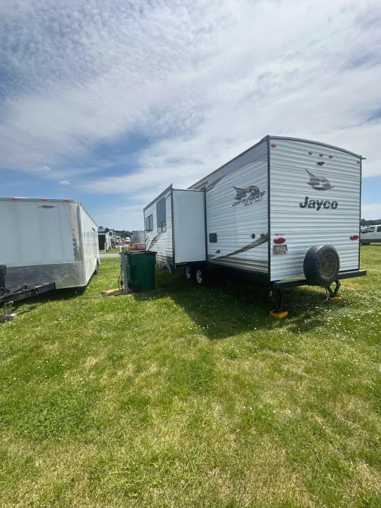 A white Jayco travel trailer RV rental, with its slide-out extended, is parked on green grass next to another trailer. Sunny day.. Jayco Jay Flight SLX 2019